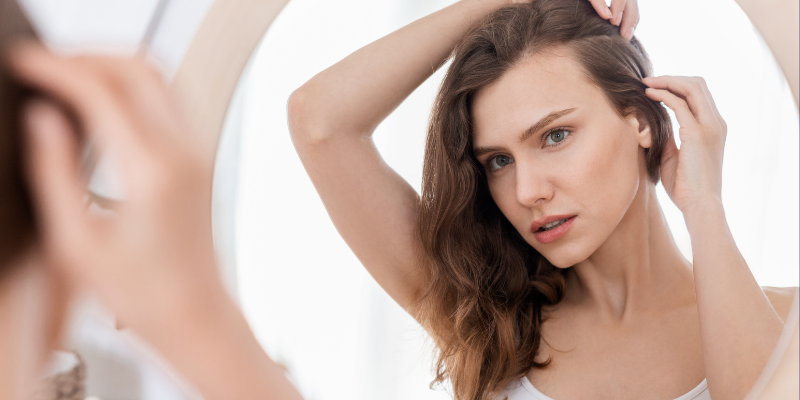 A woman examining and adjusting her wavy hair in a mirror, reflecting a moment of personal haircare management at home.