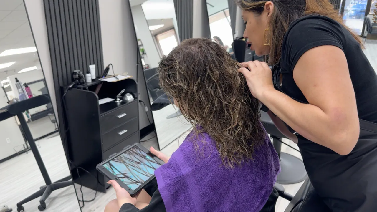Japanese Head Spa: A Path to Relaxation and Wellness 2 A professional hair stylist examines a young woman’s wet curly hair in a modern salon. The stylist uses a tablet to show the client a magnified view of her scalp condition, providing a detailed consultation before a Japanese head spa treatment. The salon is well-equipped with contemporary furniture and styling tools.