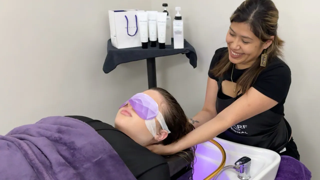Japanese Head Spa: A Path to Relaxation and Wellness 4 A professional hairstylist gently massages a client’s scalp during a Japanese head spa treatment. The client, relaxed with a lavender eye mask, reclines comfortably as the stylist uses soothing techniques to enhance scalp health. In the background, professional Milbon hair care products are displayed on a shelf, emphasizing the salon’s focus on quality treatments.